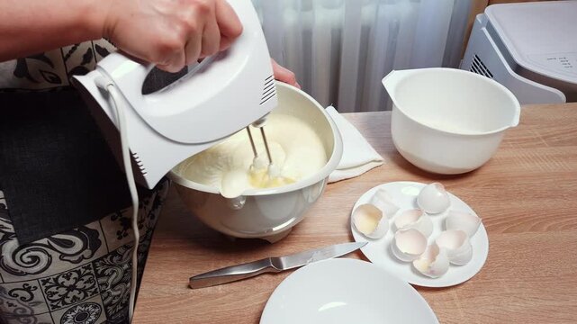 Close-up of hands using an electric mixer to blend a creamy mixture in a white bowl, with eggshells nearby on a wooden table