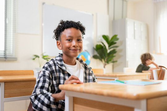 Portrait of a happy young student smiling in bright modern classroom, Cheerful elementary school boy looking at the camera during a lesson, Smart and confident diverse child sitting at desk in school