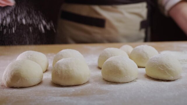 Baking flour falling on small dough balls arranged on wooden surface, cooking process.