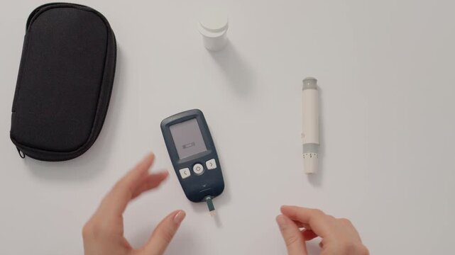A woman prepares to check her blood sugar levels using a finger pricking device. She holds the glucometer in her hands at her kitchen table.