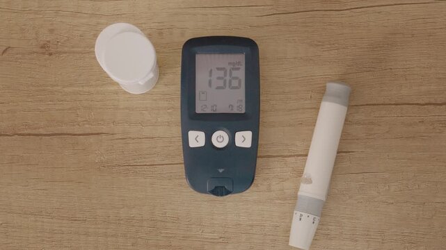 A glucometer sits on a wooden table with a reading of 136 mg dL, indicating high blood sugar. The item next to it appears to be a test strip container.