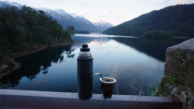A thermos and traditional mate cup rest on a railing with a serene Patagonian lake and mountains in the background. Ideal for travel content or cultural documentaries.