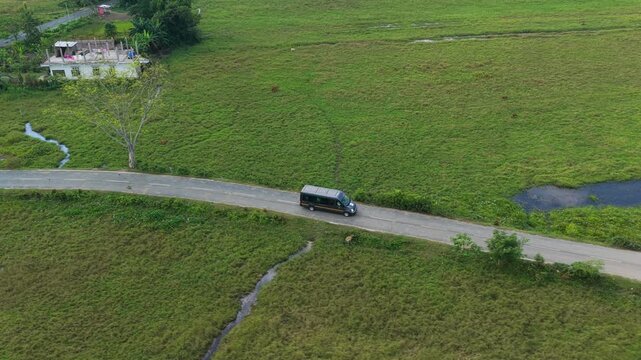 A cinematic aerial tracking shot of a passenger van traveling through lush green agricultural fields and rural landscapes in India.
