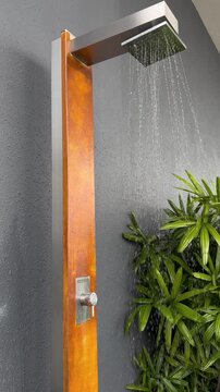 Modern outdoor rain shower with flowing water beside tropical green plants and textured concrete wall. Luxury resort spa and wellness vacation concept in tropical hotel environment.