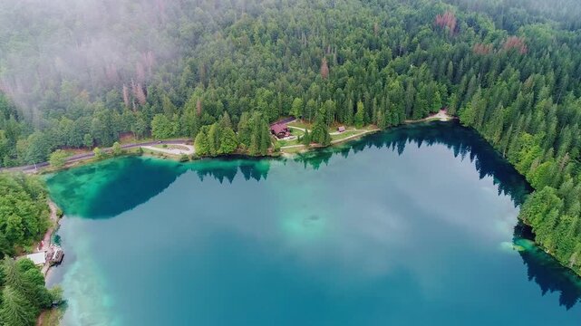 Clear alpine lake reflects forest as drone nears cozy lodge by mountain road