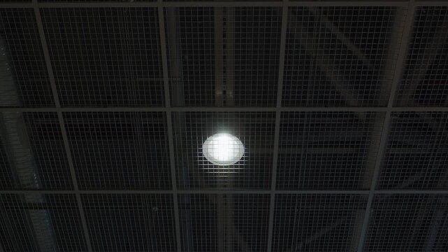 Upward view of a dark metal grid ceiling with a single bright overhead lamp, creating an industrial interior detail with geometric lines and modern utility lighting.
