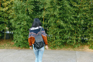 Obraz premium Back view of a woman with a backpack walking along a sidewalk among tall trees in casual autumn attire, conveying outdoor exploration and a calm park atmosphere