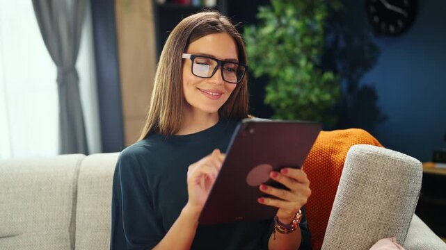 A woman sits on a couch in a living room. She interacts with a tablet and smiles while looking at the screen. The room has a modern design with plants and decor.