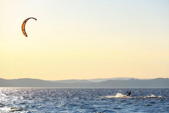 kiteboarder on lake balaton with an orange kite above evening water and the tihany peninsula with its monastery visible on the warm sunset horizon