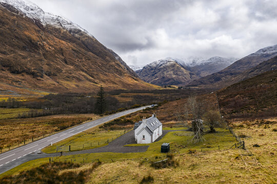 Aerial view of the iconic white cottage nestled in the dramatic Glen Coe valley with snow-capped mountains and a winding road under a cloudy sky in Scotland, United Kingdom.