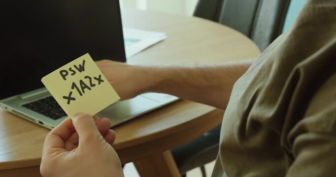 A man holds a handwritten password on a note and enters it into a laptop. Concept of cybersecurity, data privacy, weak password habits, and human error in everyday digital life.