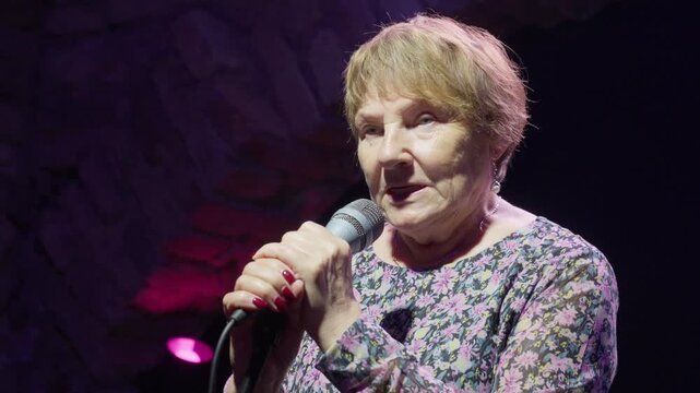Elderly woman performer talking into microphone at open mic comedy show. Senior storyteller sharing stories during public speech event at theater club stage with spotlight lighting.