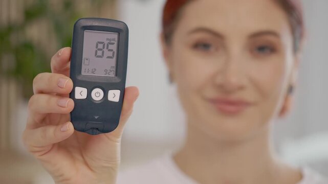 A woman holds a glucometer showing a reading of 85 mg dL. She shares her experience with managing blood sugar and diabetes at her home during the afternoon.