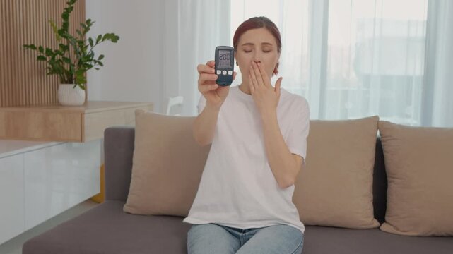 A woman sits on a couch and shows her glucometer reading of 200 mg dL, which shows a high blood sugar level that can be concerning for health.