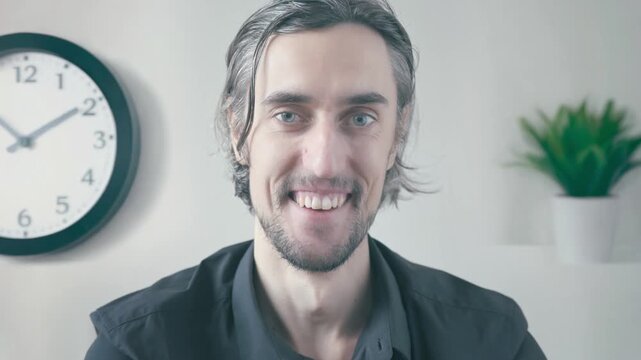 Cheerful quirky office worker with long dark hair and beard showing evolving expressions from friendly smile to serious and reassuring in simple home setting with clock and plant