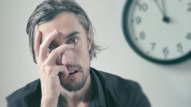 Bored office worker at work time: man long hair beard sitting by wall clock covering face with hand, nervously shifting eyes, showing anxiety, distress, frustration, and mental exhaustion indoors