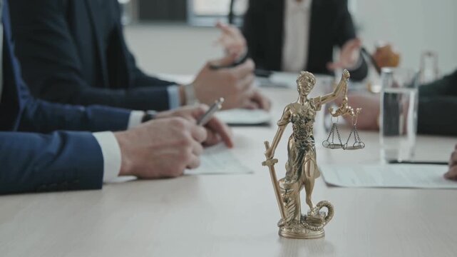 Lady Justice bronze statue on office table, legal team in background during law firm meeting, professional justice and litigation concept
