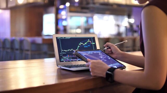 Professional stock market trader analyzing financial data on laptop and tablet, digital investment charts on screen, business woman working in cafe