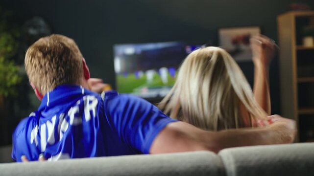Two friends sit on a couch and watch a football match on television. They cheer and react while enjoying the game. It is an evening in a cozy living room.