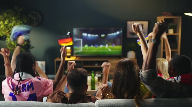 A group of friends sits on a couch in a living room and watches a soccer match on TV. They cheer and react to the game while sharing snacks and drinks.