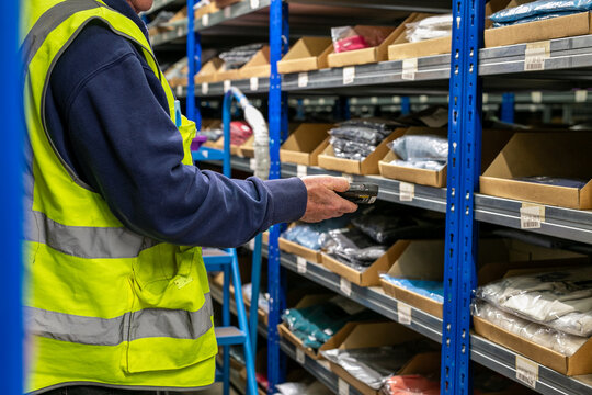 Logistics warehouse operative wearing a high-visibility yellow vest and using a wearable barcode scanner while performing inventory picking tasks in a storage aisle filled with shelved stock
