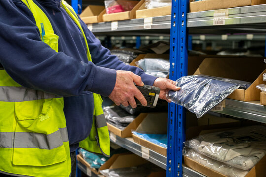 Logistics warehouse operative wearing a high-visibility yellow vest and using handheld barcode scanner to scan labels on an item while performing inventory picking tasks in an inventory storage aisle 