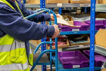 Logistics warehouse operative wearing a high-visibility yellow vest and using handheld barcode scanner to scan labels on picking transport equipment, including a shelved picking trolley, while picking © Val