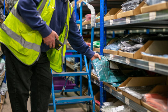Logistics warehouse operative wearing a high-visibility yellow vest and using handheld barcode scanner to scan labels on an item while performing inventory picking tasks in an inventory storage aisle 