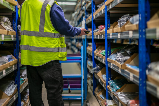 Logistics warehouse operative wearing a high-visibility yellow vest and using a wearable barcode scanner while performing inventory picking tasks in a storage aisle filled with shelved stock