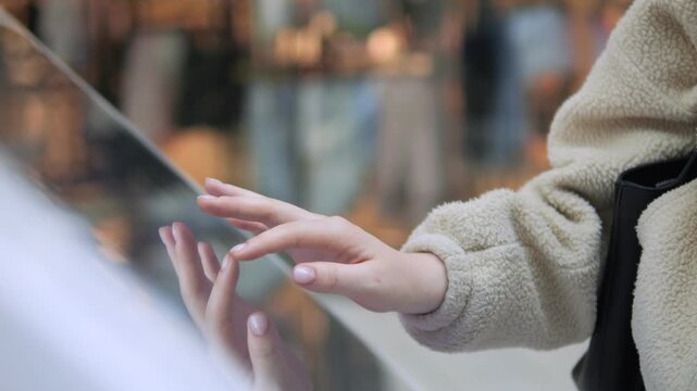 Person touches a glass surface with both hands. Fingers gently brush reflective surface. Reflection shows person's hand clearly. Background blurred, indoor setting visible. Person wears beige jacket