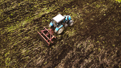 A high-angle drone shot of a blue industrial tractor pulling a red harrow across a farm field. The image captures the vehicle in mid-operation, turning over dark, rich soil and patches of green grass. © Andrey