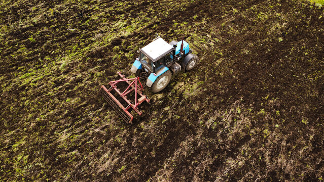 A high-angle drone shot of a blue industrial tractor pulling a red harrow across a farm field. The image captures the vehicle in mid-operation, turning over dark, rich soil and patches of green grass.