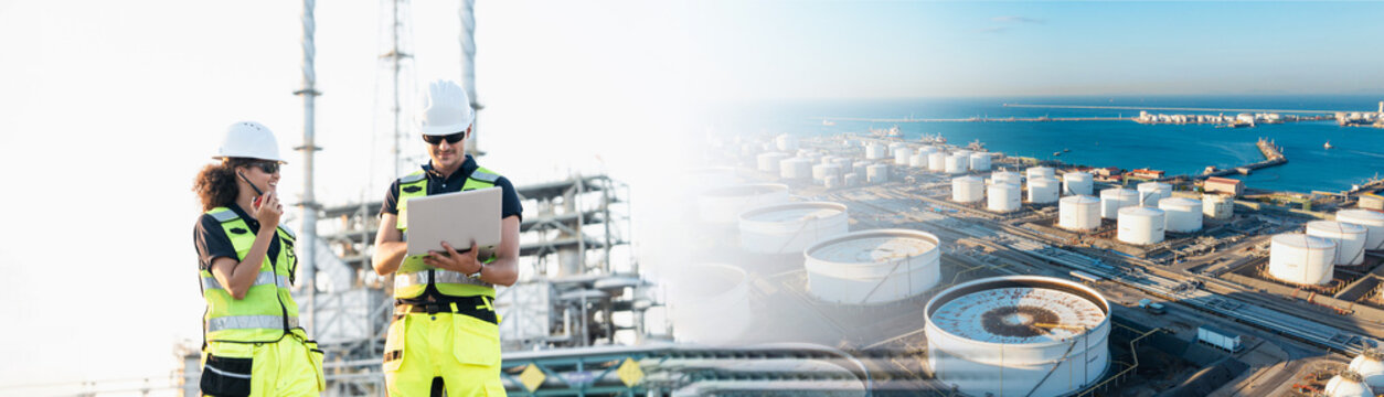 An engineer reviews data on laptop at sprawling oil refinery, examining large storage tanks and distant harbor while wearing yellow reflective vest and white hard hat on construction platform.