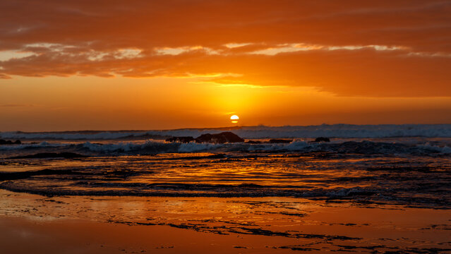 Aerial view of Hartenbos beach with waves crashing against rocks under a vibrant orange sunset sky in Mossel Bay, Western Cape, South Africa.