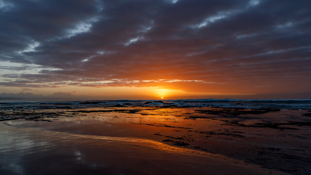 Aerial view of the sun setting over the rocky coastline of Hartenbos beach with dramatic clouds and golden reflections on the water Mossel Bay, Western Cape, South Africa.