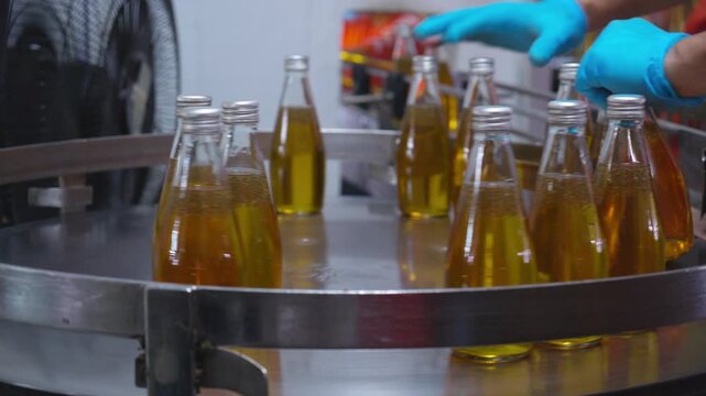Factory worker hand checking glass bottles with yellow liquid fruit juice beverage moving on metal conveyor belt, automated manufacturing production line facility, food processing plant.