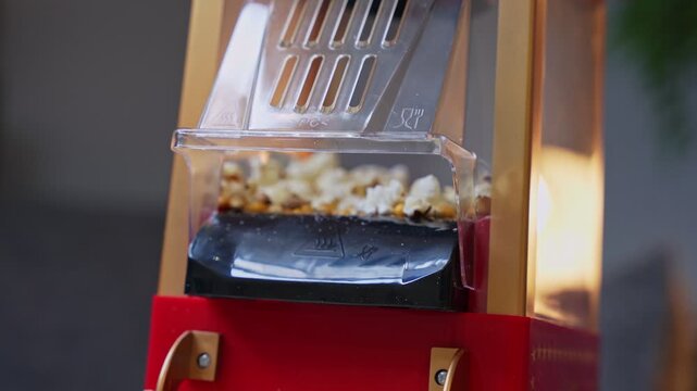 Fresh popcorn popping and falling from a red vintage-style machine indoors. High-speed motion highlights the snack preparation for a home cinema or movie night event.