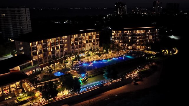 A night aerial shot of the Dusit Thani Mactan Cebu resort in the Philippines, highlighting the illuminated pool area and surrounding buildings reflecting on the water.