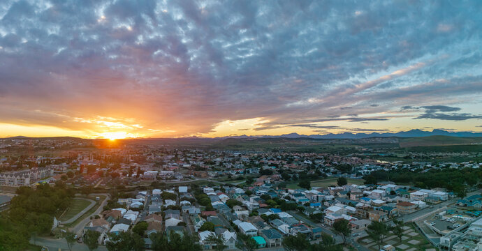 Aerial view of Hartenbos residential area with rooftops and streets under a dramatic sunset sky and mountain range in Mossel Bay, Western Cape, South Africa.