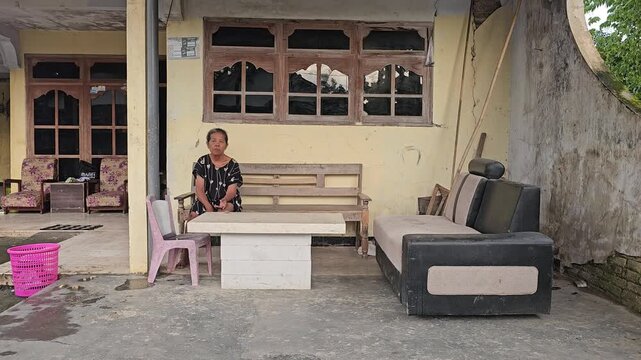 Elderly Asian woman resting on a wooden bench on the front porch of a modest rural house surrounded by various furniture
