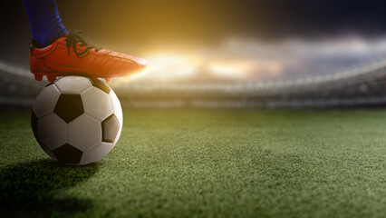 A close-up shot of a soccer player's foot in a red cleat resting on a ball on a green turf field inside a blurred stadium at sunset or under spotlights © Leo Lintang