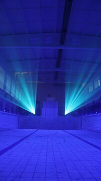 Symmetrical turquoise light beams fill a blue-lit empty indoor pool hall with tiled floor, high ceiling, haze, distant platform, and copy space