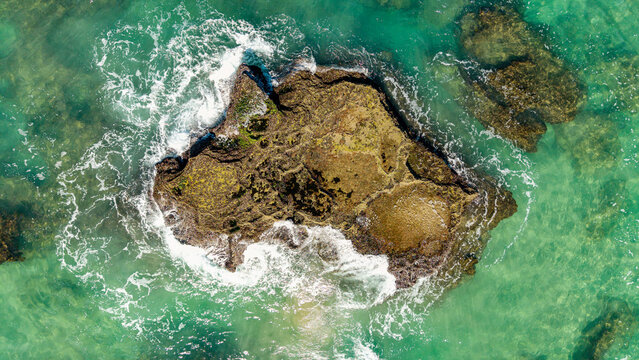 Aerial view of a rugged rocky reef surrounded by crystal clear turquoise ocean water and crashing white waves near Hartenbos Mossel Bay, Western Cape, South Africa.