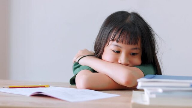 Close up selective focus of a tired Asian kid girl resting her head on the desk. Stressed and exhausted from studying too much homework in bright daylight. Education burnout and overwork concept.