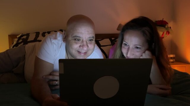 Pareja de mediana edad acostada boca abajo en la cama riendo frente al port&aacute;til
