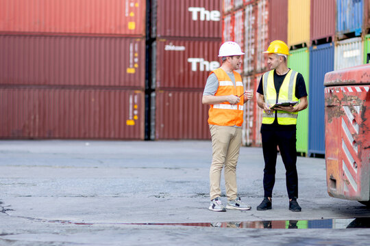 Two workers with safety vest and helmet working together at logistic shipping cargo container yard. Male worker writing on clipboard during discussing with his engineer foreman who holding worky talky