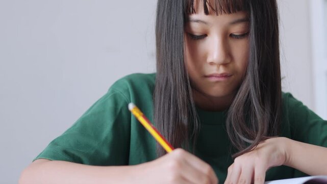 Close up selective focus of a concentrated Asian kid, a young Thai girl or Chinese student, diligently doing homework and writing with a pencil in a notebook. Education, learning, and study concept.