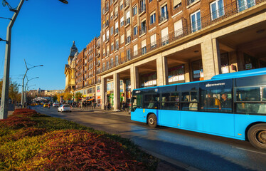 Obraz premium Blue public transport bus driving on a city street past historic European architecture and residential buildings in Budapest, Hungary, during a bright sunny day.