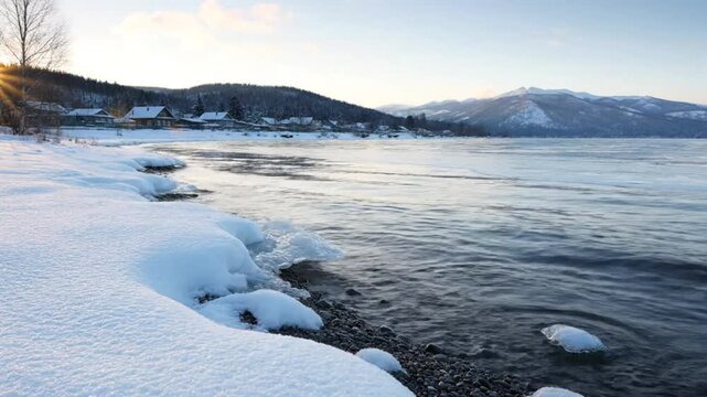Scenic winter shoreline with snow-covered beach, gently lapping waves, and a distant village under a clear blue sky