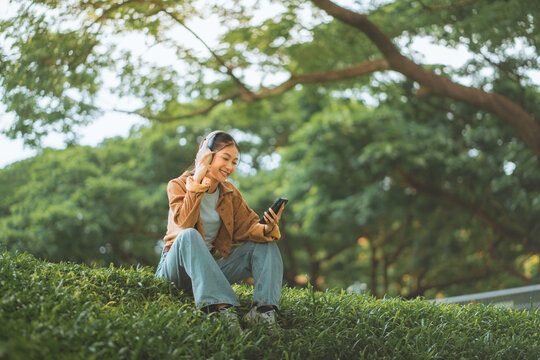 Happy young Asian woman sitting on grass in a green park, wearing headphones and using her smartphone to listen to music. Peaceful female student relaxing outdoors under a tree. digital lifestyle
